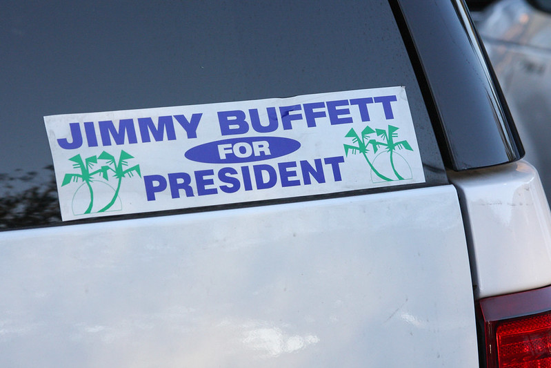 A bumper sticker on a car that says "Jimmy Buffett for President" in bold purple text, flanked by green palm tree illustrations.
