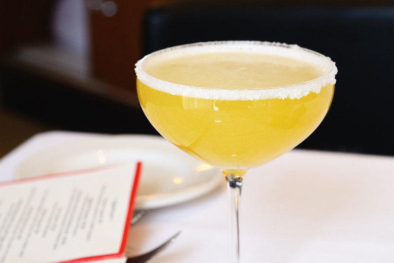 A golden margarita in a coupe glass with a sugared rim, set on a white tablecloth with a menu and plate in the background.
