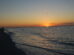 Sunset on a beach with gentle waves, silhouetted birds flying, and people walking along the shoreline. A distant city skyline is visible on the horizon under the orange and golden glow of the setting sun.