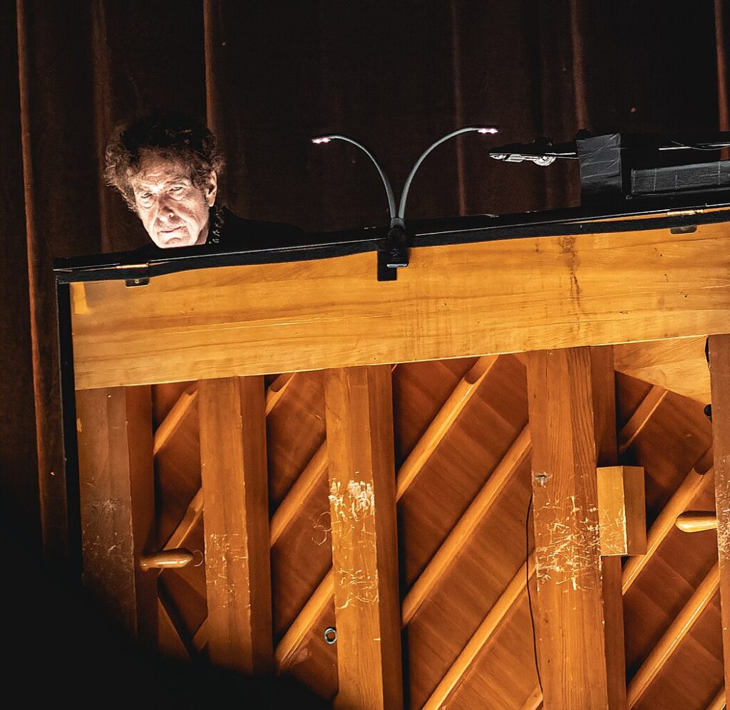 Bob Dylan, with an intense expression, peers over a grand piano during a live performance, with dramatic lighting and a dark stage background.