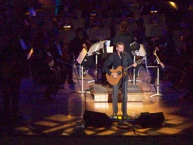 Kenny Loggins performing with a guitar on a brightly lit stage, accompanied by the Boston Pops Orchestra in the background.