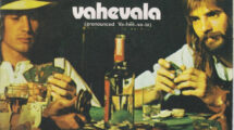 "Cover of the single 'Vahevala' by Kenny Loggins and Jim Messina, showing the duo sitting at a poker table with cards, drinks, and a bottle of wine." Description: "This is the cover art for the single 'Vahevala' by Kenny Loggins and Jim Messina. It depicts the duo seated at a poker table surrounded by cards, glasses, and a bottle of wine, capturing a relaxed and casual vibe. The text prominently displays the title 'Vahevala' with a pronunciation guide (Va-hee-va-la) and also includes the title of the B-side track, 'Same Old Wine.'"
