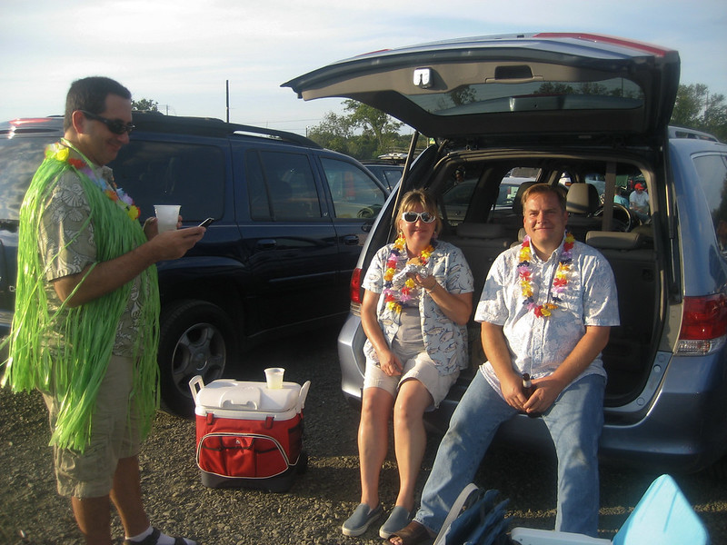 Three people dressed in tropical shirts and leis relax at a tailgate, with a cooler and a minivan in the background.