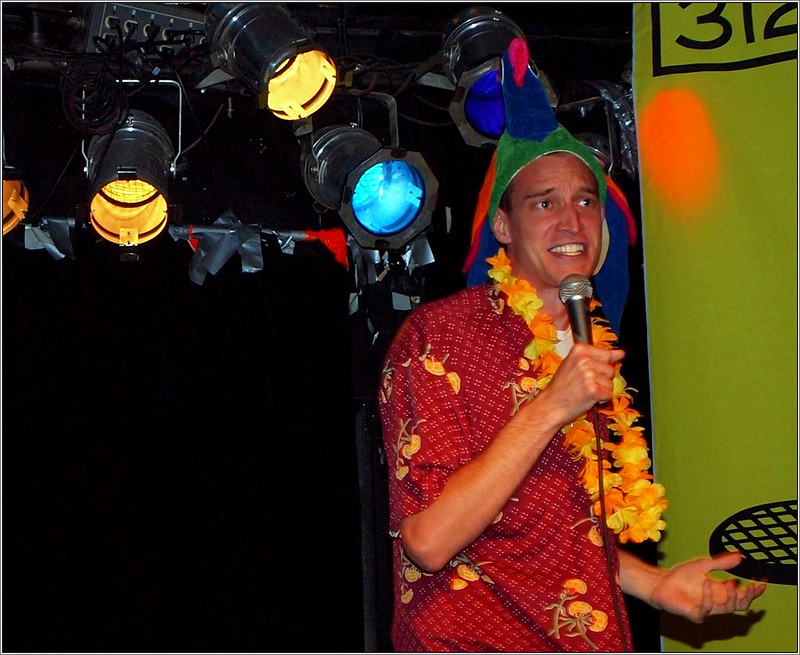 A man wearing a colorful parrot hat, a Hawaiian shirt, and a yellow lei sings into a microphone under stage lights. Maybe he's a member of The Official Jimmy Buffett Fan Club?