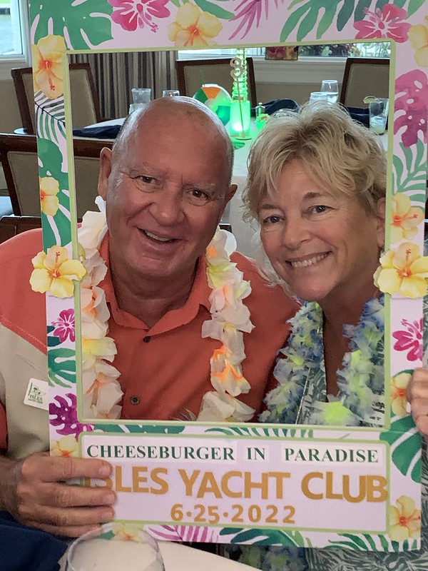 "Two people wearing leis smile while holding a tropical-themed photo frame reading 'Cheeseburger in Paradise' at Naples Yacht Club, June 25, 2022.
