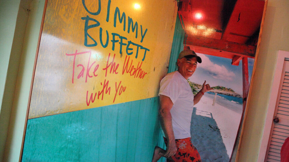 Jimmy Buffett smiling in front of a colorful tropical poster reading 'Take the Weather With You,' wearing a white shirt and red swim trunks with a beach in the background.