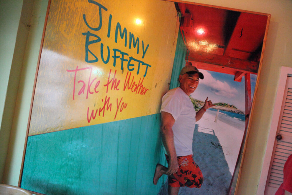 Jimmy Buffett smiling in front of a colorful tropical poster reading 'Take the Weather With You,' wearing a white shirt and red swim trunks with a beach in the background.