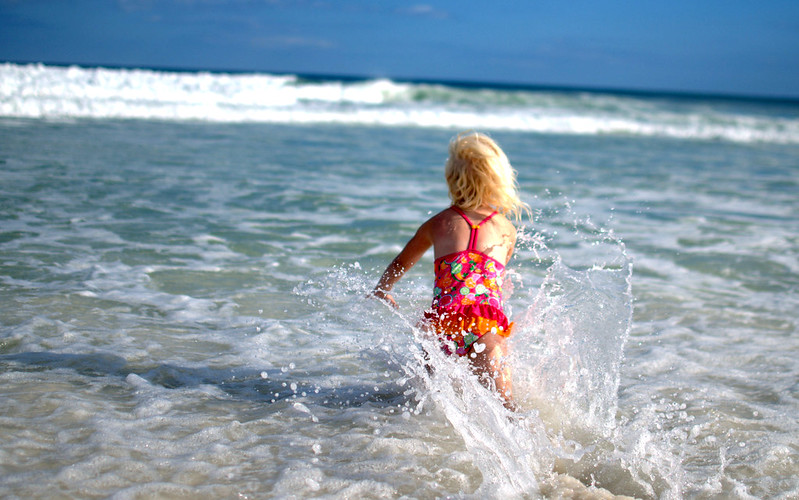 A young child in a colorful swimsuit runs into the waves, splashing water under a clear blue sky at the beach