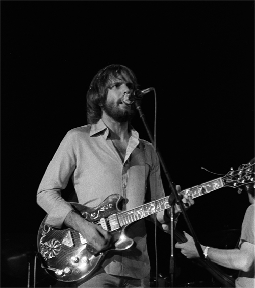 Bob Weir playing guitar and singing into a microphone during a live performance, wearing a light-colored shirt and focused expression.