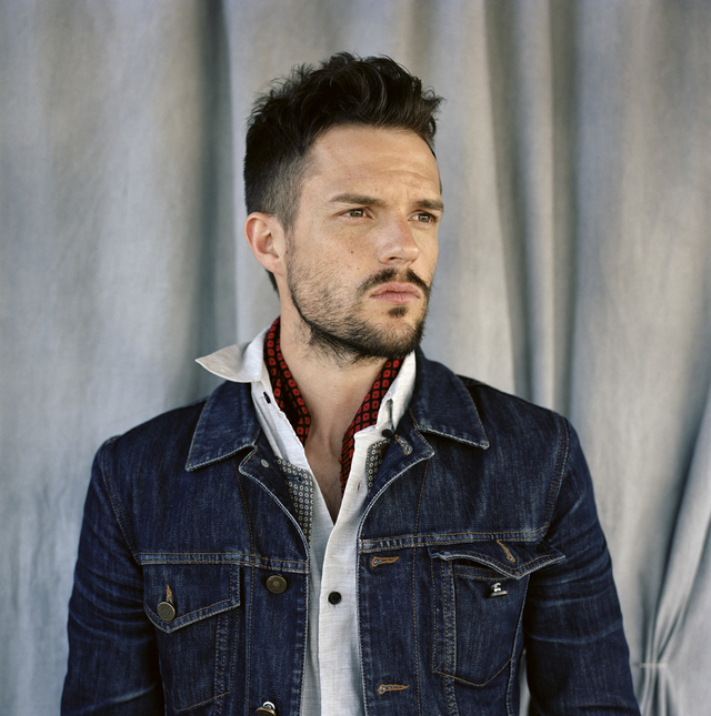 Brandon Flowers, lead singer of The Killers, wearing a denim jacket and white shirt, posing in front of a neutral backdrop.