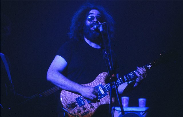 Jerry Garcia playing guitar and singing into a microphone during a live Grateful Dead performance, with his iconic glasses and black shirt.