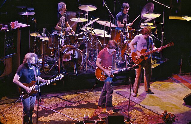 The Grateful Dead performing on stage with Jerry Garcia on guitar, Bob Weir, Phil Lesh, and two drummers, featuring a classic rock setup.