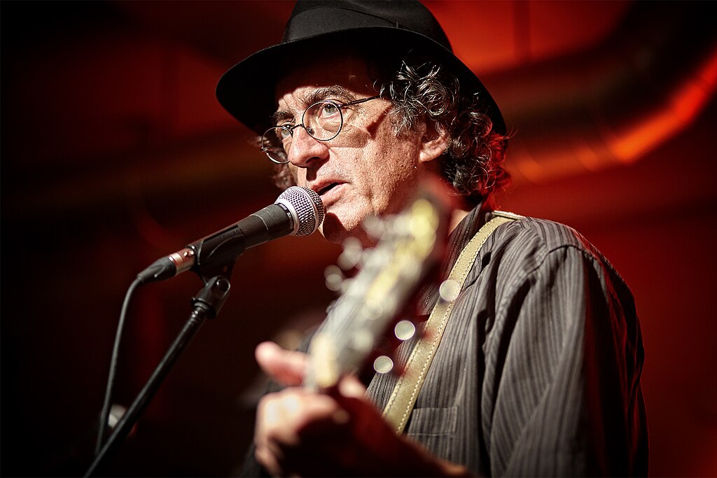James McMurtry, wearing a black hat and glasses, performs on stage with a microphone and guitar, lit by warm red lighting.