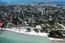 Aerial view of Key West, Florida, with turquoise waters, a sandy shoreline, tennis courts, and a dense collection of homes and buildings stretching inland.