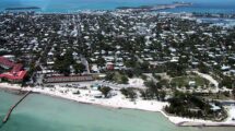 Aerial view of Key West, Florida, with turquoise waters, a sandy shoreline, tennis courts, and a dense collection of homes and buildings stretching inland.