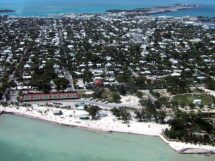 Aerial view of Key West, Florida, with turquoise waters, a sandy shoreline, tennis courts, and a dense collection of homes and buildings stretching inland.