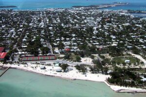Aerial view of Key West, Florida, with turquoise waters, a sandy shoreline, tennis courts, and a dense collection of homes and buildings stretching inland.