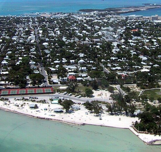 Aerial view of Key West, Florida, with turquoise waters, a sandy shoreline, tennis courts, and a dense collection of homes and buildings stretching inland.