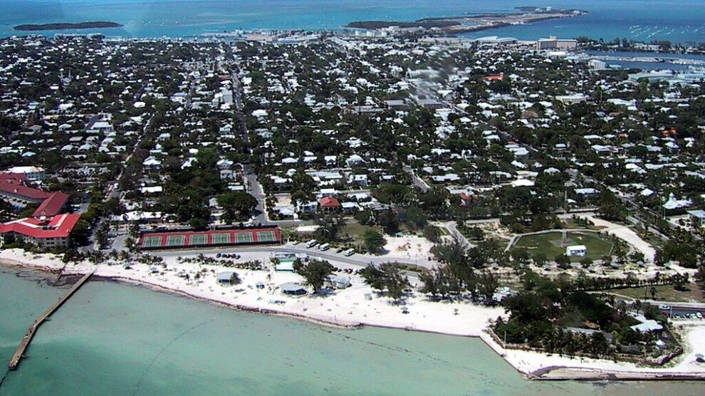 Aerial view of Key West, Florida, with turquoise waters, a sandy shoreline, tennis courts, and a dense collection of homes and buildings stretching inland.