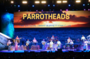 A live band performs on stage with a tropical-themed background featuring a sunset over the ocean and the text "Parrotheads, The Official Jimmy Buffett Fan Club