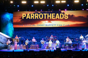 A live band performs on stage with a tropical-themed background featuring a sunset over the ocean and the text "Parrotheads, The Official Jimmy Buffett Fan Club