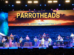 A live band performs on stage with a tropical-themed background featuring a sunset over the ocean and the text "Parrotheads, The Official Jimmy Buffett Fan Club