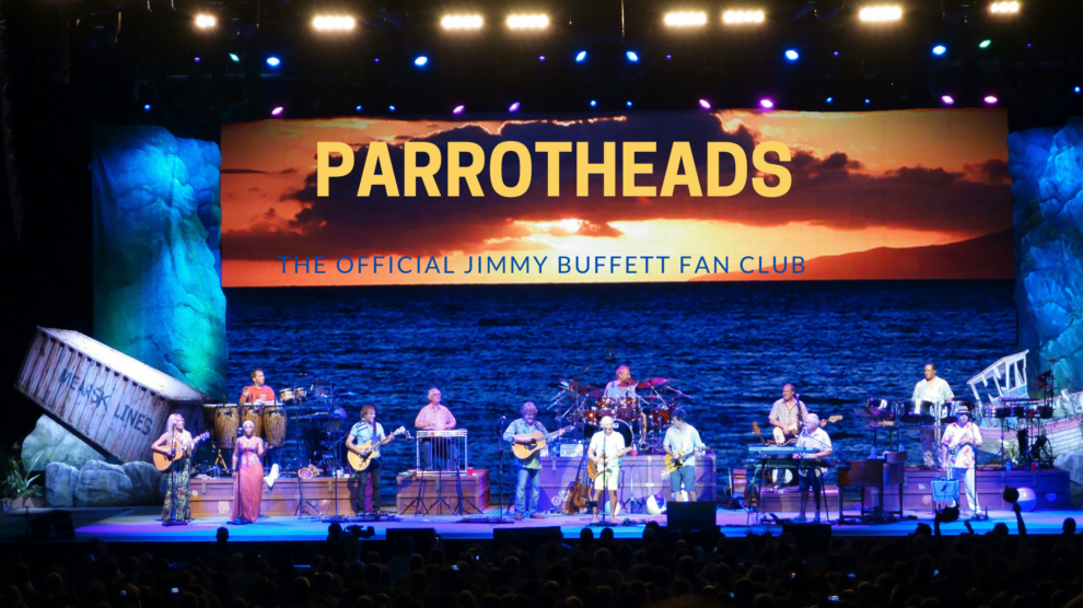 A live band performs on stage with a tropical-themed background featuring a sunset over the ocean and the text "Parrotheads, The Official Jimmy Buffett Fan Club
