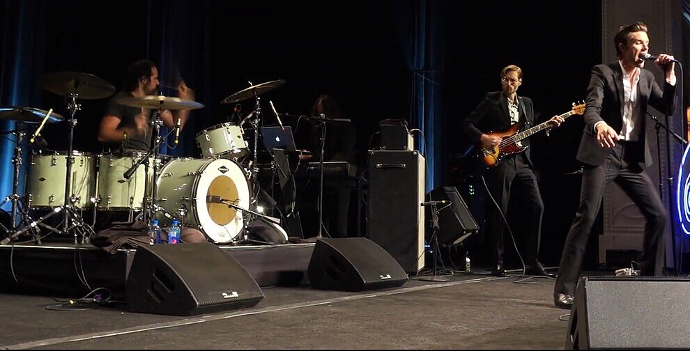 The Killers performing live in Las Vegas, featuring Brandon Flowers singing, Ronnie Vannucci Jr. on drums, and Mark Stoermer on bass.