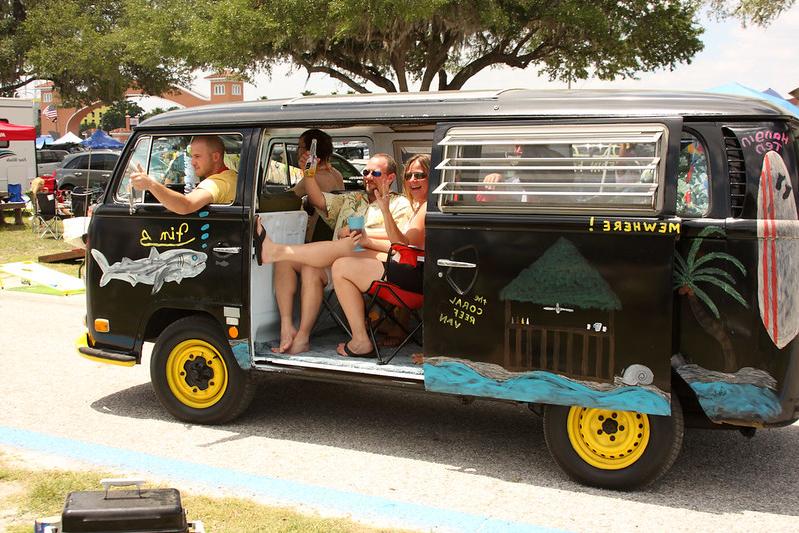 A black vintage van decorated with tropical art, including palm trees and ocean waves, with smiling people inside waving and enjoying drinks.