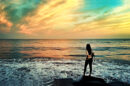 A woman standing on a rock by the ocean at sunset, looking at the waves with a colorful sky in the background.