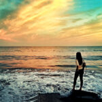 A woman standing on a rock by the ocean at sunset, looking at the waves with a colorful sky in the background.