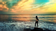 A woman standing on a rock by the ocean at sunset, looking at the waves with a colorful sky in the background.