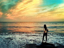 A woman standing on a rock by the ocean at sunset, looking at the waves with a colorful sky in the background.