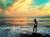 A woman standing on a rock by the ocean at sunset, looking at the waves with a colorful sky in the background.