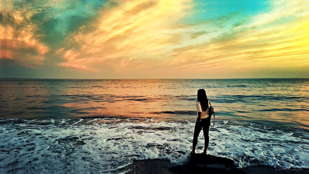 A woman standing on a rock by the ocean at sunset, looking at the waves with a colorful sky in the background.
