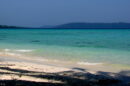 A tropical beach with gentle waves washing onto the white sandy shore, with clear turquoise waters stretching towards a distant tree-covered island under a hazy blue sky.
