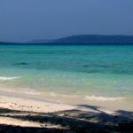 A tropical beach with gentle waves washing onto the white sandy shore, with clear turquoise waters stretching towards a distant tree-covered island under a hazy blue sky.