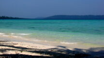 A tropical beach with gentle waves washing onto the white sandy shore, with clear turquoise waters stretching towards a distant tree-covered island under a hazy blue sky.