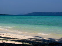 A tropical beach with gentle waves washing onto the white sandy shore, with clear turquoise waters stretching towards a distant tree-covered island under a hazy blue sky.