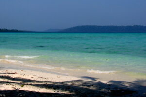 A tropical beach with gentle waves washing onto the white sandy shore, with clear turquoise waters stretching towards a distant tree-covered island under a hazy blue sky.