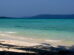 A tropical beach with gentle waves washing onto the white sandy shore, with clear turquoise waters stretching towards a distant tree-covered island under a hazy blue sky.