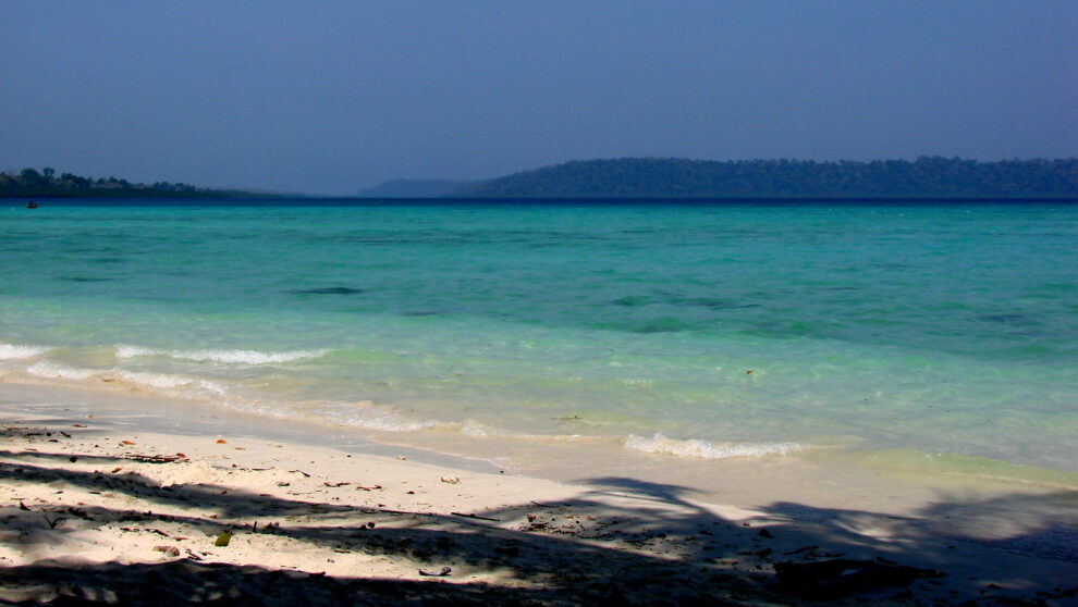 A tropical beach with gentle waves washing onto the white sandy shore, with clear turquoise waters stretching towards a distant tree-covered island under a hazy blue sky.