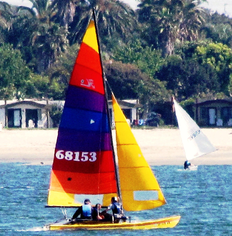 A multicolored sailboat with three sailors navigating near a beach with palm trees and small cabins in the background.