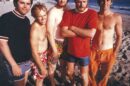 The Beach Boys standing on a beach, dressed in colorful swimwear, smiling and relaxed, representing their California surf culture image.