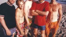 The Beach Boys standing on a beach, dressed in colorful swimwear, smiling and relaxed, representing their California surf culture image.