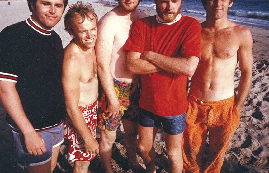 The Beach Boys standing on a beach, dressed in colorful swimwear, smiling and relaxed, representing their California surf culture image.