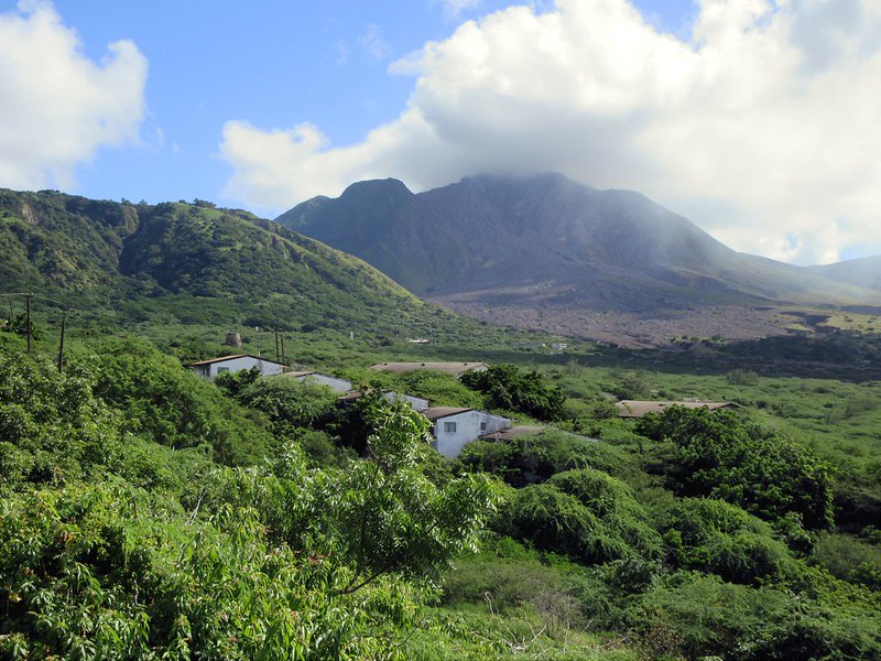 A landscape featuring Soufrière Hills Volcano on Montserrat, with a partially cloud-covered peak. In the foreground, green vegetation surrounds abandoned buildings from the former town of Plymouth, which was evacuated due to volcanic eruptions in the 1990s.