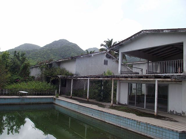 A dilapidated building with a collapsed roof and rusting supports, part of the ruins of AIR Studios Montserrat. In front of the structure is a pool filled with green water, surrounded by cracked tiles. Lush green mountains rise in the background, contrasting with the abandoned state of the former recording studio.