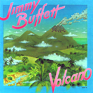 Jimmy Buffett's Volcano album cover featuring illustrated green mountains, a smoking volcano, tropical foliage, and a blue sky with scattered clouds. The album title "Volcano" is displayed in pink and white letters, with "Jimmy Buffett" prominently written at the top in a bold, stylized font. The vinyl cover is positioned in front of a shelf filled with records.