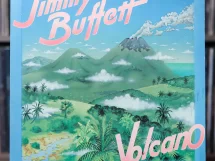 Jimmy Buffett's Volcano album cover featuring illustrated green mountains, a smoking volcano, tropical foliage, and a blue sky with scattered clouds. The album title "Volcano" is displayed in pink and white letters, with "Jimmy Buffett" prominently written at the top in a bold, stylized font. The vinyl cover is positioned in front of a shelf filled with records. Let me know if you’d like any modifications! 🎶🌋